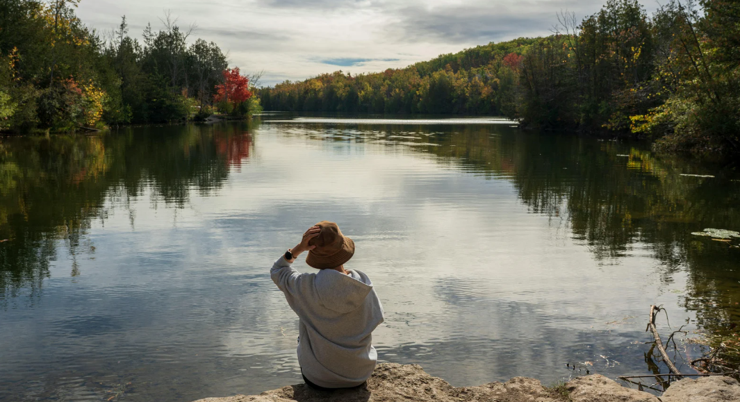 A person wearing a hat and sweatshirt sits on rocks, looking out over a body of water lined with trees on both sides.