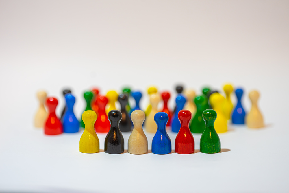 Six small colorful game pieces shaped like people stand in a line with many others in a group behind them