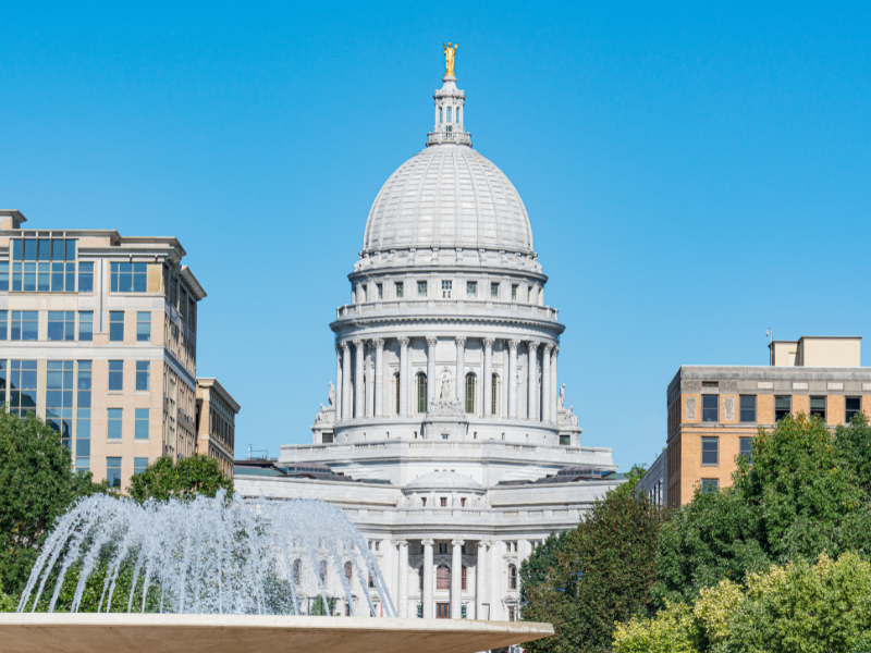 Tan buildings and green treetops surround the gray Wisconsin State Capitol. Water runs in a small fountain in the left corner.